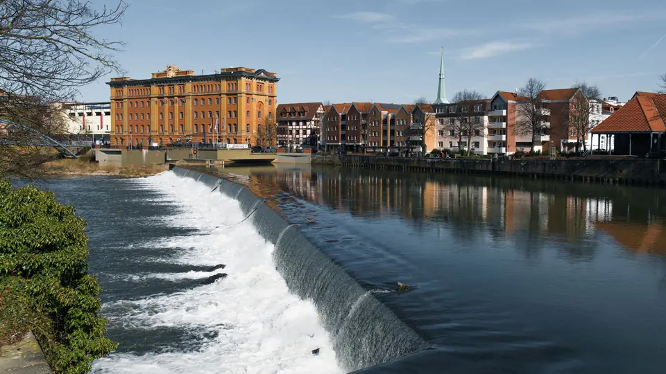 Ruhige Szene am Hamelner Flussufer mit Wasserfall und historischen Gebäuden.