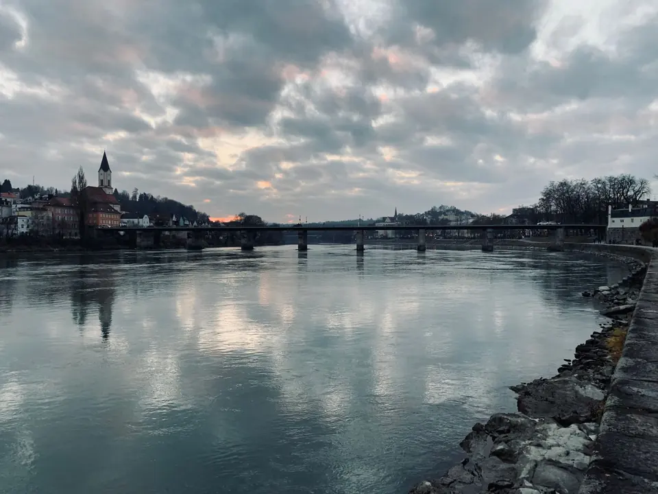 Eine ruhige Ansicht der Brücke in Passau, Deutschland, die sich im ruhigen Fluss bei Dämmerung mit einem bewölkten Himmel spiegelt.